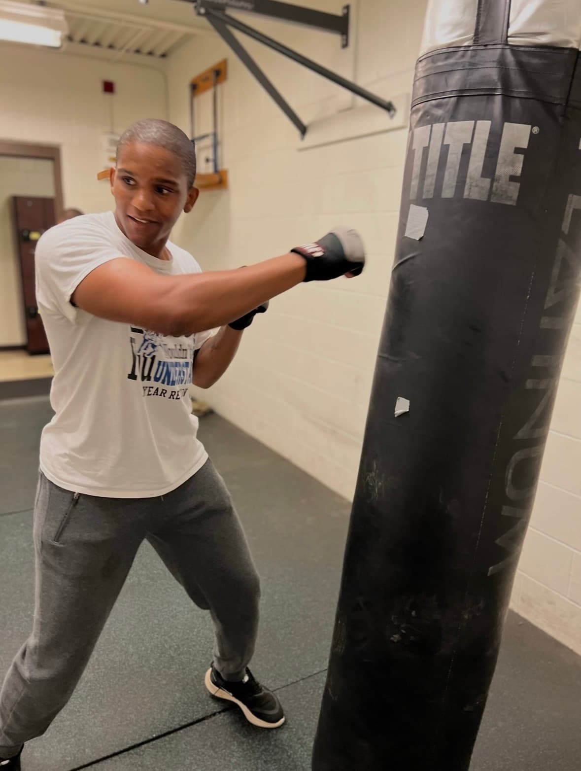A young man wearing boxing gloves punches a black heavy bag in a gym.