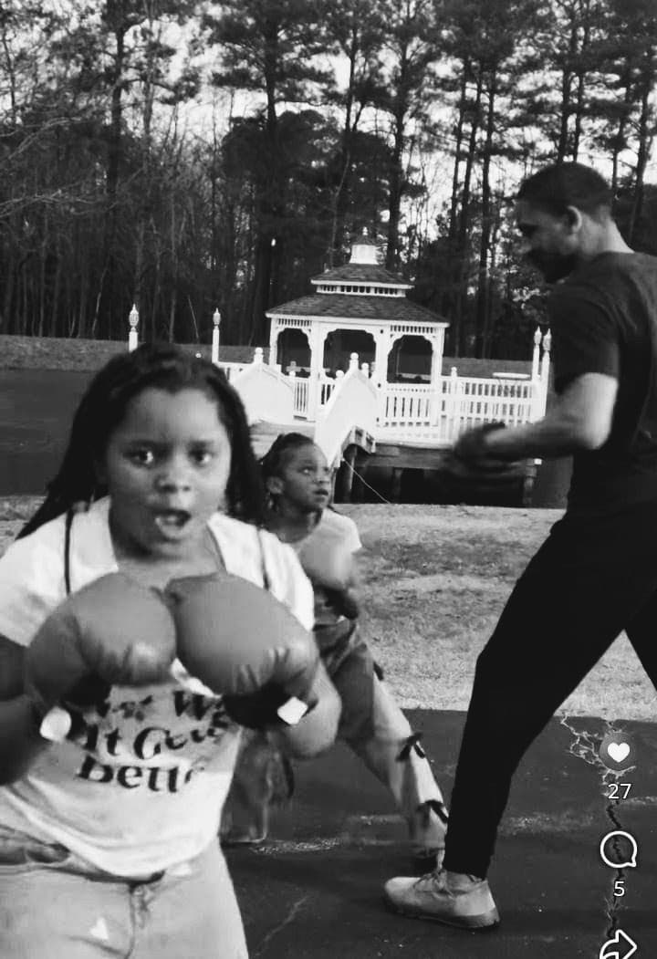 Young girl wearing boxing gloves practices with a man outdoors near a white gazebo.