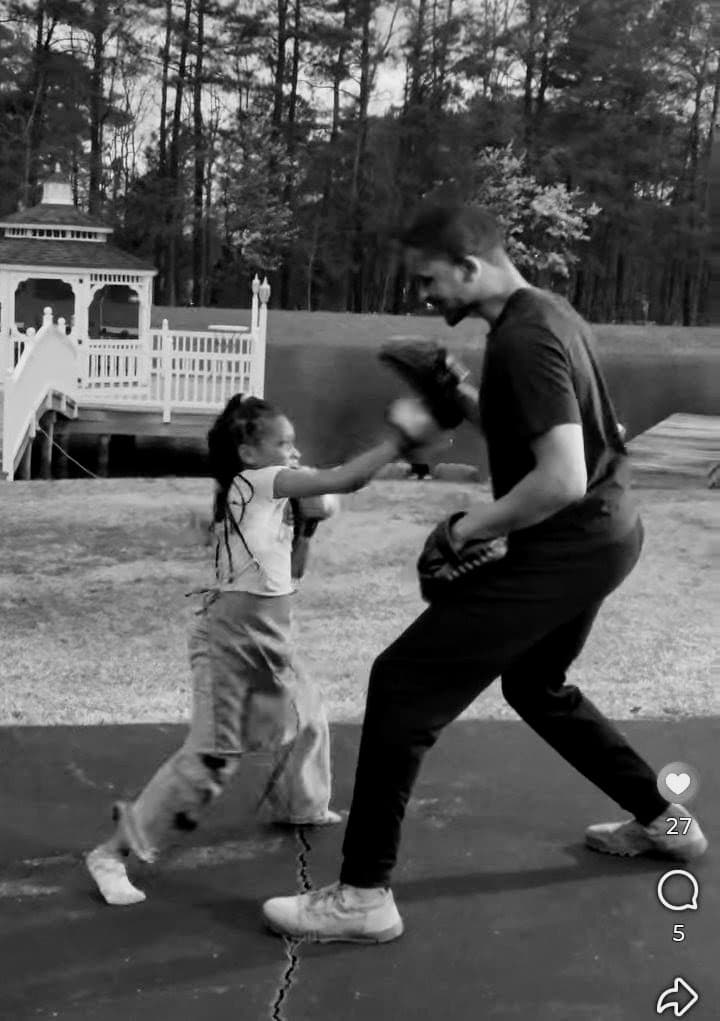 A young girl practices boxing with a man holding focus mitts in an outdoor setting.